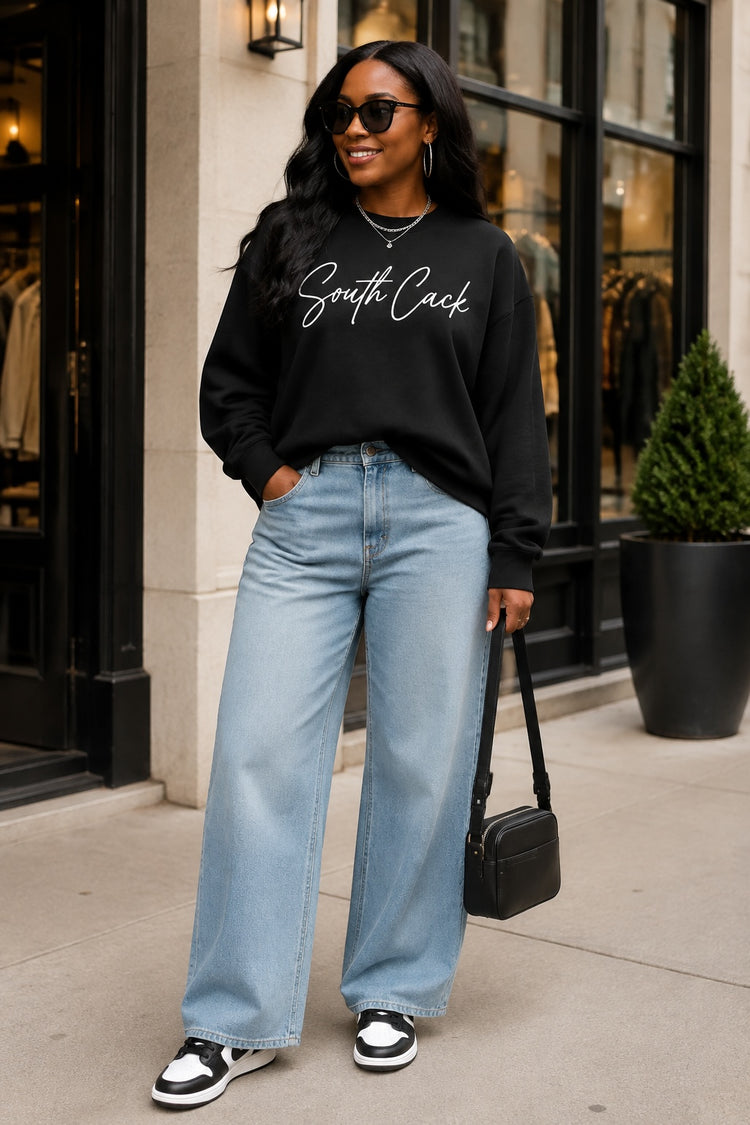 Woman wearing a black saysomethingteez South Cack crewneck sweatshirt with light wash wide-leg jeans and sneakers.